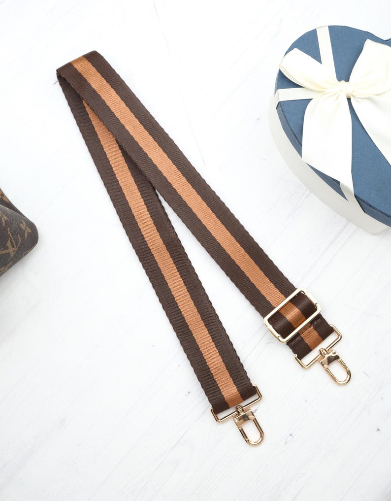 Op de afbeelding: Een bruin en tan gestreepte tasriem met goudkleurige metalen hardware. De riem heeft een geweven textuur en is ontworpen om aan een tas te worden bevestigd. De riem wordt getoond op een witte ondergrond.