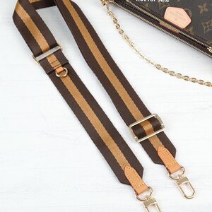 May include: Brown and tan striped handbag strap with gold-tone hardware. The strap features a brown base with a tan stripe down the center. The strap has gold-tone metal clasps and adjustable buckles. The bag is not for sale.