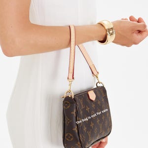 Puede incluir: Bolso de mano marr&oacute;n y beige con monograma y correa dorada. El bolso tiene forma rectangular y cierre de cremallera. El texto en el bolso dice "The bag is not for sale". Una pulsera dorada en la mu&ntilde;eca.