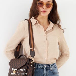 Op de afbeelding: Bruin en zwart geruite handtas met een brede, gestreepte schouderriem. De tas heeft een klepsluiting en goudkleurige hardware. De tekst op de tas luidt "The bag is not for sale".