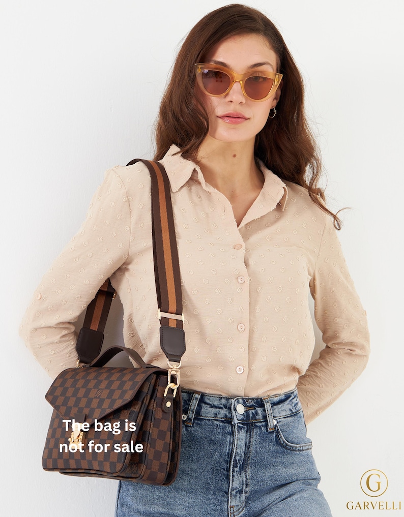 Op de afbeelding: Bruin en zwart geruite handtas met een brede, gestreepte schouderriem. De tas heeft een klepsluiting en goudkleurige hardware. De tekst op de tas luidt "The bag is not for sale".
