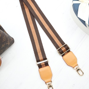 Op de afbeelding: Een bruin en beige gestreepte tasriem met goudkleurige hardware. De riem heeft een geweven ontwerp met twee beige strepen in het midden. De uiteinden hebben beige leren patches en goudkleurige sluitingen voor bevestiging.