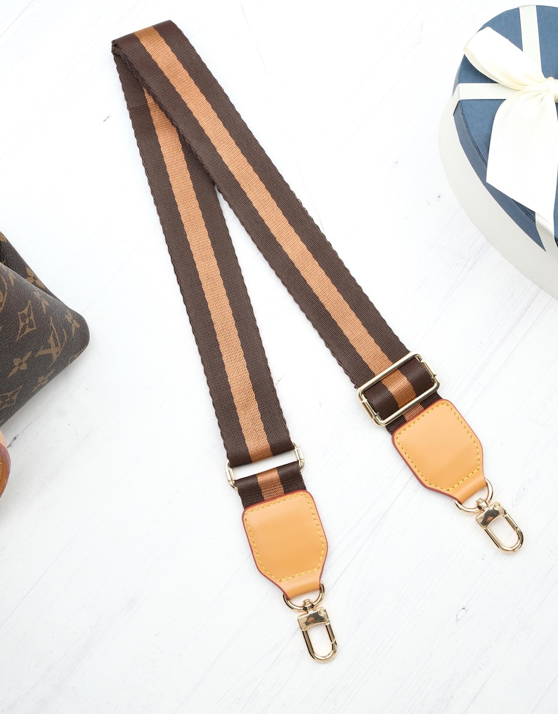 Op de afbeelding: Een bruin en beige gestreepte tasriem met goudkleurige hardware. De riem heeft een geweven ontwerp met twee beige strepen in het midden. De uiteinden hebben beige leren patches en goudkleurige sluitingen voor bevestiging.