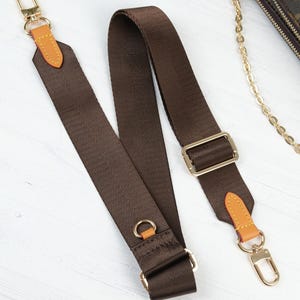May include: A brown nylon purse strap with gold-colored metal hardware. The strap has tan leather accents and a gold-colored clasp. The strap is designed to be attached to a purse or bag.