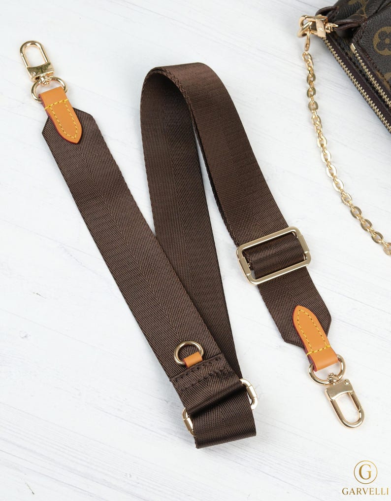 May include: A brown nylon purse strap with gold-colored metal hardware. The strap has tan leather accents and a gold-colored clasp. The strap is designed to be attached to a purse or bag.