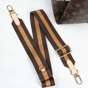 May include: A brown and tan striped bag strap with gold-colored hardware. The strap has a rectangular buckle and two gold-colored clasps. The text "The bag is not for sale" is visible in the background.