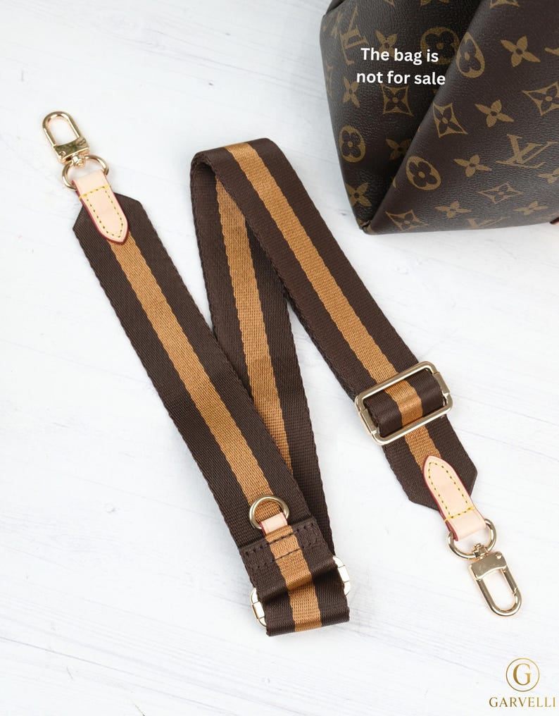 May include: A brown and tan striped bag strap with gold-colored hardware. The strap has a rectangular buckle and two gold-colored clasps. The text "The bag is not for sale" is visible in the background.