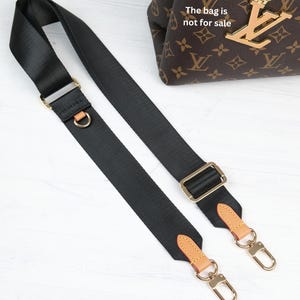 May include: Black nylon bag strap with gold-tone hardware and tan leather accents. The strap features adjustable buckles and swivel clasps for easy attachment. The bag in the background is not for sale.