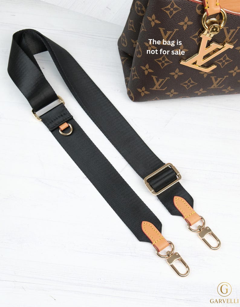 May include: Black nylon bag strap with gold-tone hardware and tan leather accents. The strap features adjustable buckles and swivel clasps for easy attachment. The bag in the background is not for sale.