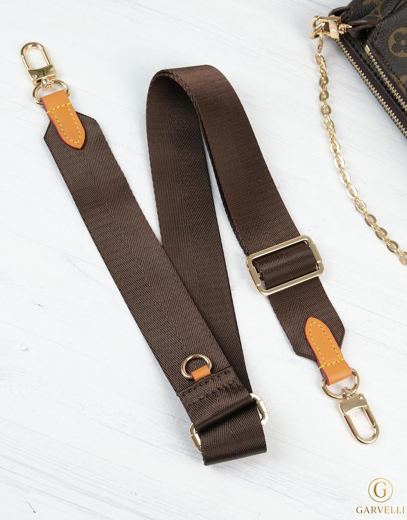 May include: A brown nylon purse strap with gold-colored metal hardware. The strap has tan leather accents and a gold-colored clasp on each end. The strap is designed to be adjustable and is shown on a white wooden surface.