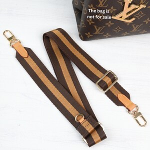 May include: Brown and tan striped bag strap with gold-tone hardware. The strap has leather accents and metal clasps. The bag in the background has a brown monogram pattern and a gold charm. The text on the bag says "The bag is not for sale."