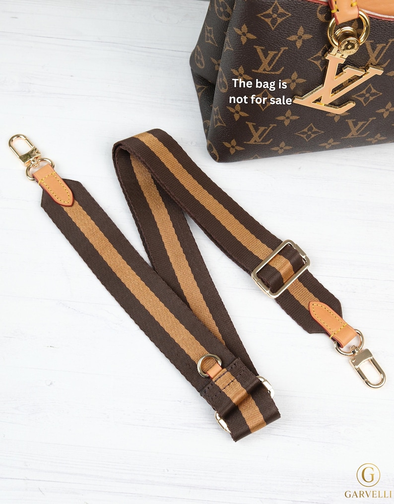 May include: Brown and tan striped bag strap with gold-tone hardware. The strap has leather accents and metal clasps. The bag in the background has a brown monogram pattern and a gold charm. The text on the bag says "The bag is not for sale."