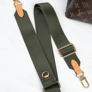 May include: Olive green canvas bag strap with gold-tone hardware and tan leather accents. The strap features adjustable buckles and swivel clasps for easy attachment to a bag. The strap is designed for use with a handbag.