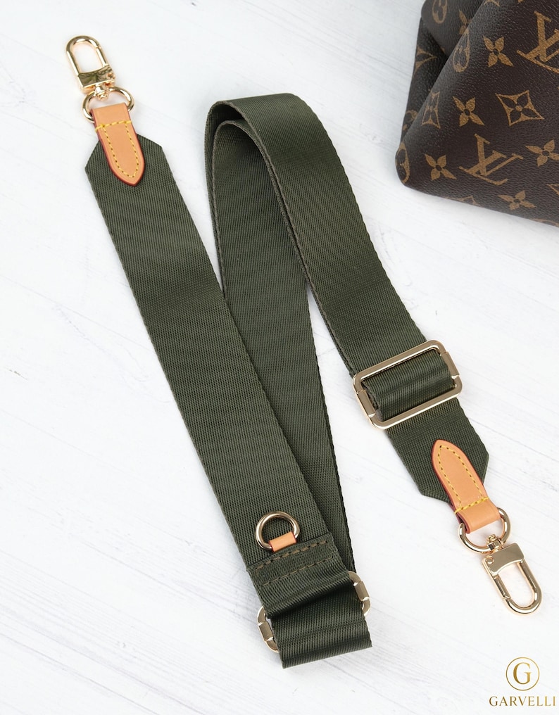 May include: Olive green canvas bag strap with gold-tone hardware and tan leather accents. The strap features adjustable buckles and swivel clasps for easy attachment to a bag. The strap is designed for use with a handbag.
