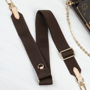 May include: A brown canvas shoulder strap with gold-tone hardware, including clasps, rings, and a rectangular buckle. The strap has light tan leather accents at the ends. A portion of a brown and tan patterned bag is visible in the background.