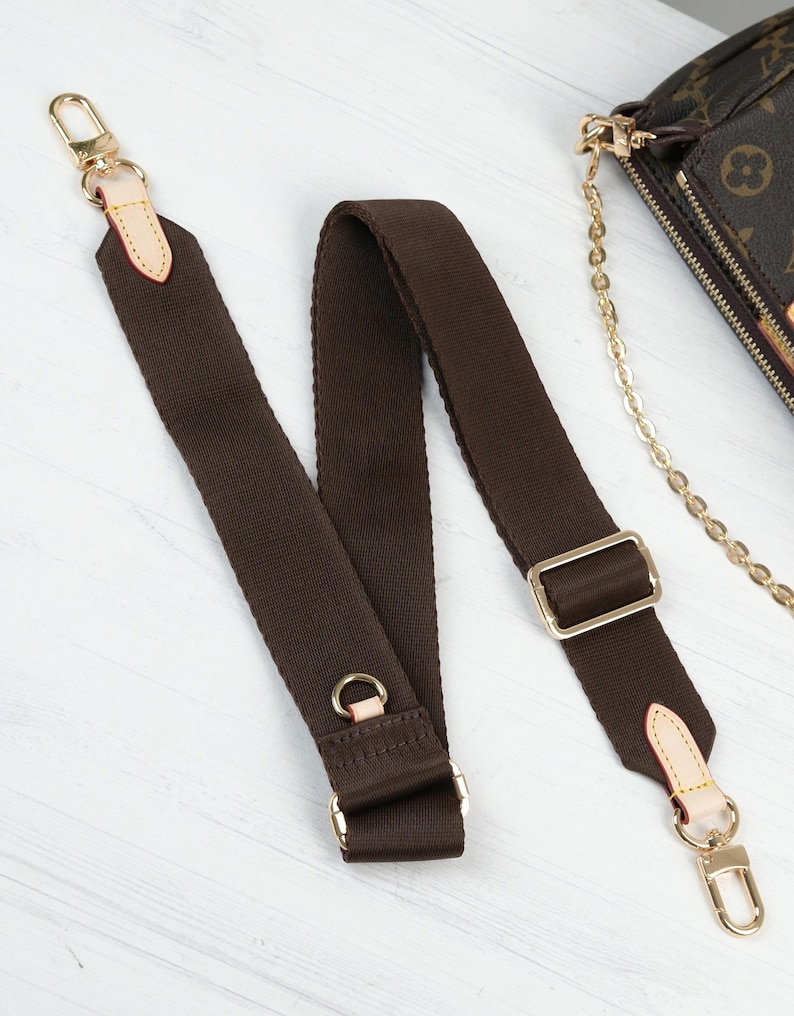 May include: A brown canvas shoulder strap with gold-tone hardware, including clasps, rings, and a rectangular buckle. The strap has light tan leather accents at the ends. A portion of a brown and tan patterned bag is visible in the background.