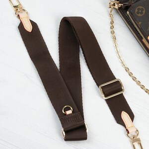 May include: A brown canvas shoulder strap with gold-tone hardware, including clasps, rings, and a rectangular buckle. The strap has light tan leather accents at the ends. A portion of a brown and tan patterned bag is visible in the background.
