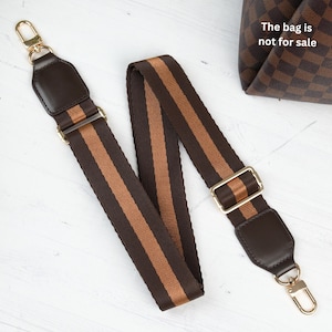 Op de afbeelding: Een bruin en tan gestreepte tasriem met goudkleurige hardware. De riem heeft aan elk uiteinde een lederen accent en een goudkleurige gesp. De tasriem wordt getoond op een wit oppervlak.