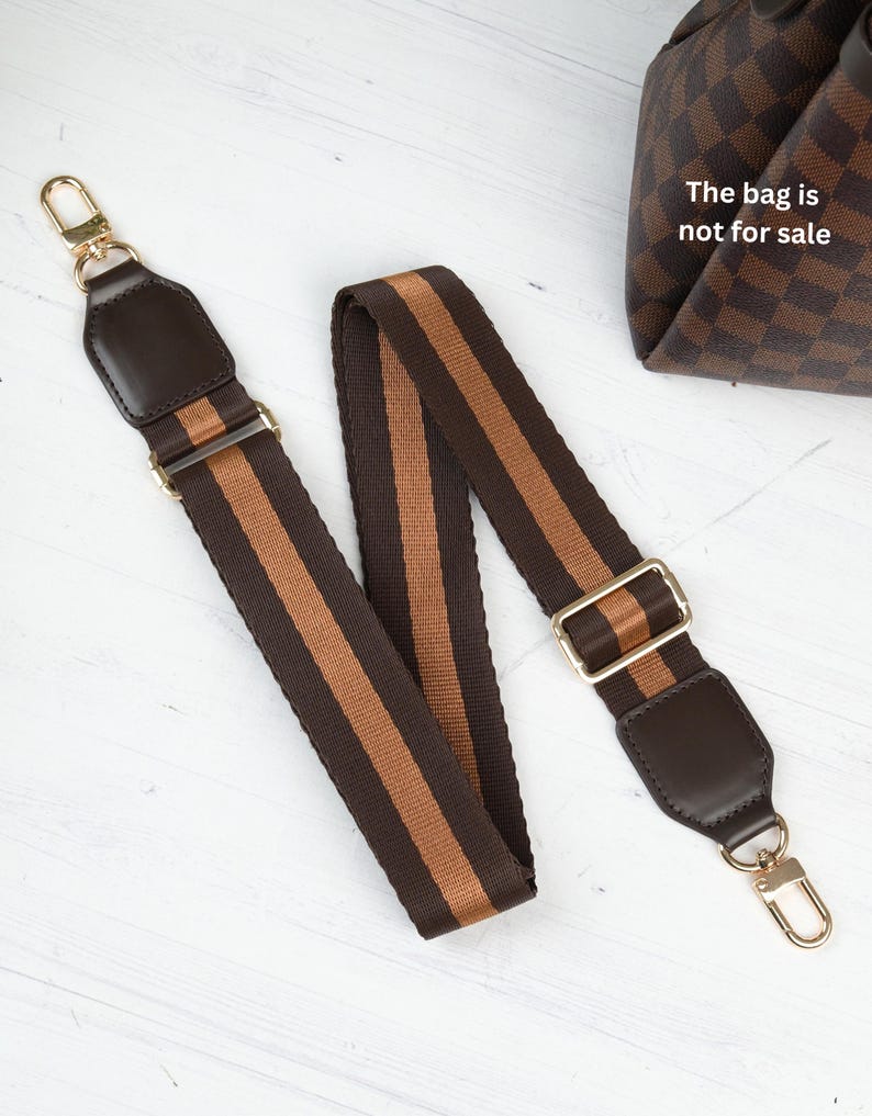 Op de afbeelding: Een bruin en tan gestreepte tasriem met goudkleurige hardware. De riem heeft aan elk uiteinde een lederen accent en een goudkleurige gesp. De tasriem wordt getoond op een wit oppervlak.