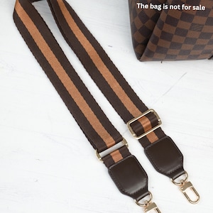 Op de afbeelding: Bruin en tan gestreepte tasriem met goudkleurige hardware. De riem heeft donkerbruine lederen accenten en goudkleurige sluitingen voor eenvoudige bevestiging. De tas op de achtergrond is niet te koop.