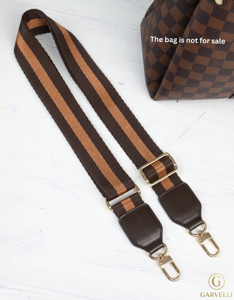 Op de afbeelding: Bruin en tan gestreepte tasriem met goudkleurige hardware. De riem heeft donkerbruine lederen accenten en goudkleurige sluitingen voor eenvoudige bevestiging. De tas op de achtergrond is niet te koop.