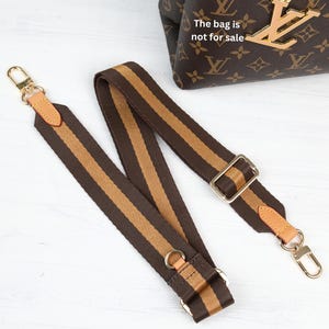 May include: Brown and tan striped bag strap with gold-tone hardware. The strap has leather accents and metal clasps. The bag in the background has a brown monogram pattern. The text on the bag says "The bag is not for sale."