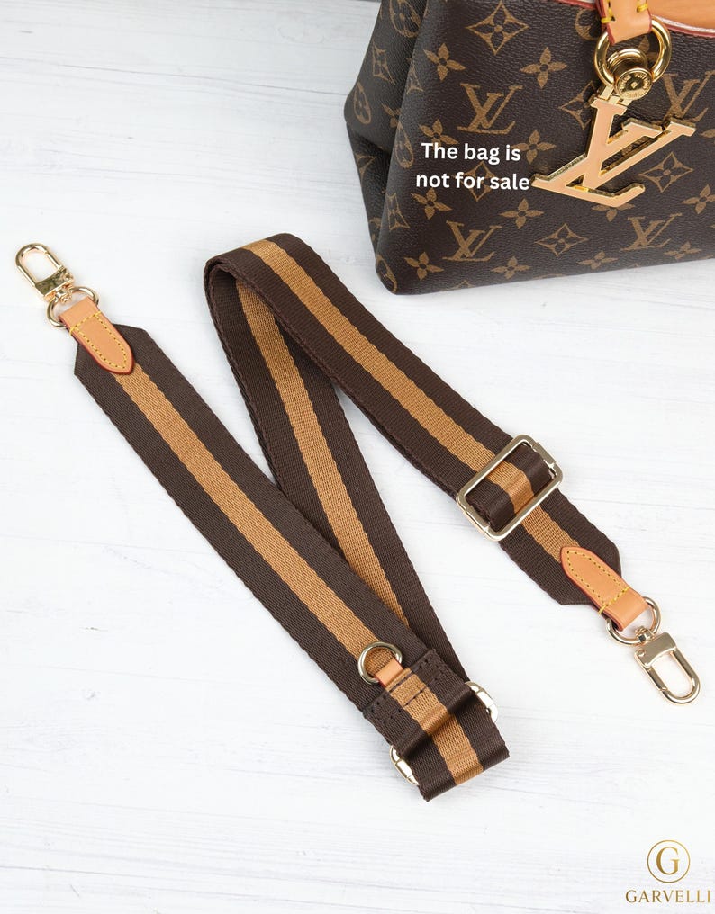 May include: Brown and tan striped bag strap with gold-tone hardware. The strap has leather accents and metal clasps. The bag in the background has a brown monogram pattern. The text on the bag says "The bag is not for sale."