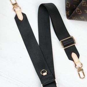 May include: Black adjustable bag strap with gold-tone hardware, including clasps, rings, and a buckle. The strap is made of a durable, woven material. A portion of a brown patterned bag is visible in the background.