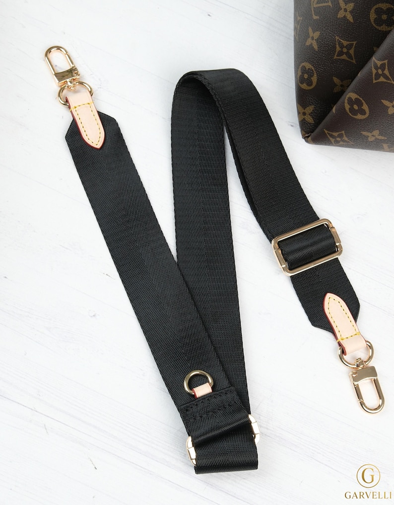May include: Black adjustable bag strap with gold-tone hardware, including clasps, rings, and a buckle. The strap is made of a durable, woven material. A portion of a brown patterned bag is visible in the background.