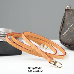 Puede incluir: Una correa de cuero vachetta de plena flor de color marr&oacute;n claro con bordes rojos y cierres met&aacute;licos dorados. La correa mide 1 cm de ancho. El texto "Full-Grain Vachetta Leather" es visible.