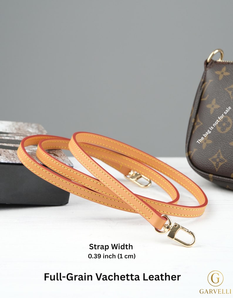 Puede incluir: Una correa de cuero vachetta de plena flor de color marr&oacute;n claro con bordes rojos y cierres met&aacute;licos dorados. La correa mide 1 cm de ancho. El texto "Full-Grain Vachetta Leather" es visible.
