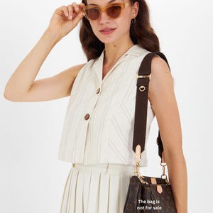 May include: A woman wearing light brown sunglasses and a cream-colored outfit, holding the sunglasses. A brown monogrammed handbag with a brown strap hangs from her shoulder. The text on the bag reads, "The bag is not for sale."