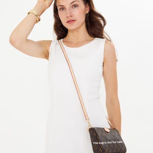 Puede incluir: Un bolso bandolera marr&oacute;n con un estampado de monograma y una correa clara. El bolso lo lleva una persona con un vestido blanco. El texto en el bolso dice "La bolsa no est&aacute; a la venta."