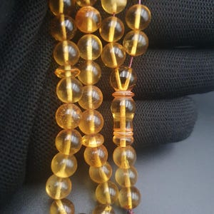 Puede incluir: Primer plano de un collar de cuentas de oración de ámbar. Las cuentas son de color amarillo dorado translúcido, ensartadas con un fino cordón rojo. La borla es negra.