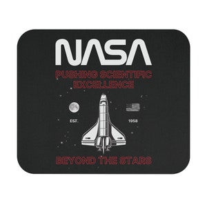 Puede incluir: Un ratón de ordenador negro con un logotipo blanco de la NASA, el texto "Pushing Scientific Excellence", una lanzadera espacial, el texto "Est. 1958", y el texto "Beyond the Stars".