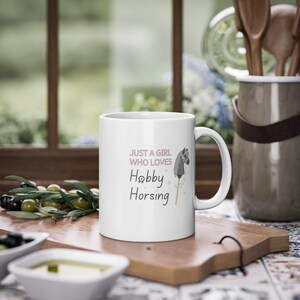 May include: White ceramic mug with a pink and gray design. The mug features a stick horse with a gray horse head and the text "Just a girl who loves Hobby Horsing".