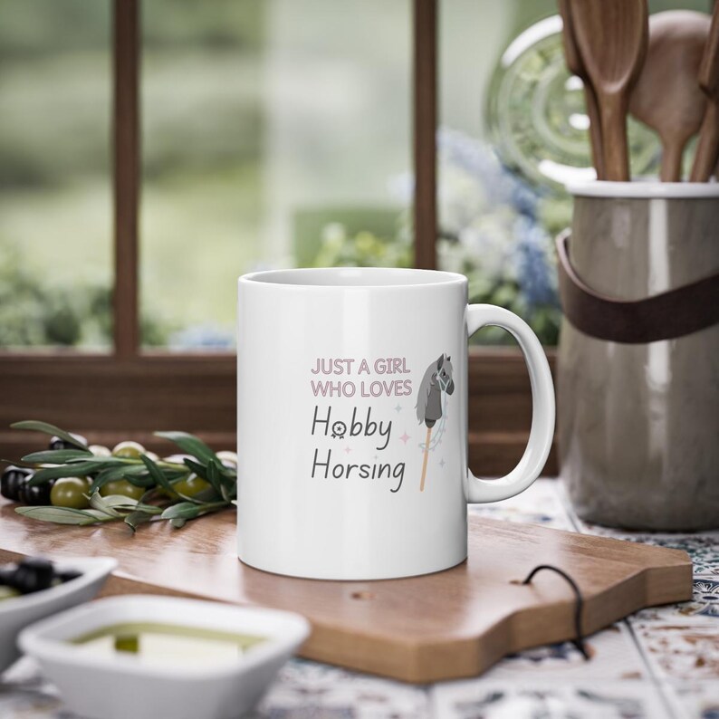 May include: White ceramic mug with a pink and gray design. The mug features a stick horse with a gray horse head and the text "Just a girl who loves Hobby Horsing".
