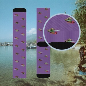 Puede incluir: Un par de calcetines morados con un patrón repetido de una criatura verde sosteniendo un sable de luz. Los calcetines tienen un puño y una punta negros.