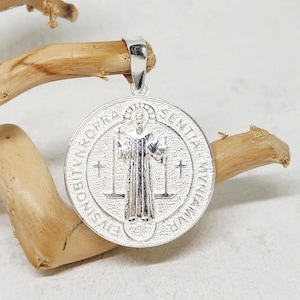 May include: A silver pendant with a detailed depiction of a religious figure, surrounded by text. The pendant is round with a loop at the top for a chain. The background is a light color, and the pendant is resting on a piece of wood.