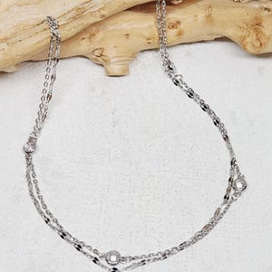 May include: A silver-toned double-strand necklace featuring delicate metallic chains with small, evenly spaced circular clear stones set in metal bezels. The necklace has a reflective finish and a layered design that creates a subtle, elegant appearance.