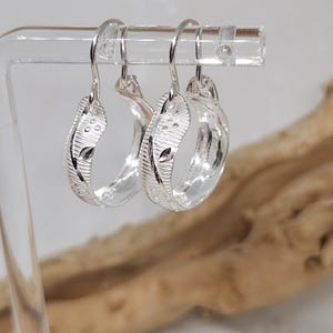 May include: A pair of silver hoop earrings with a textured design. The earrings are hanging on a display stand.
