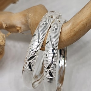 May include: Two silver bangles with an intricate floral design. The bangles are stacked on top of each other and are resting on a piece of wood.