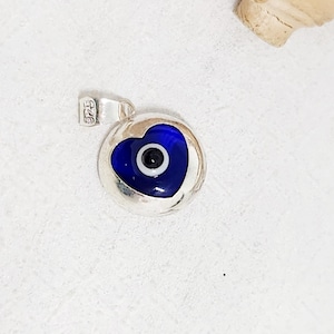 May include: A silver pendant featuring a blue heart-shaped evil eye design with a black center. The pendant has a polished finish and a small loop for a chain. The background is a light, textured surface.