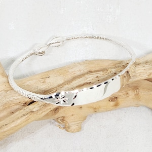 Op de afbeelding: Een zilverkleurige armband met een rechthoekig middelpunt en een bloemmotief. De armband heeft een delicate, patroonband en een sluiting. De sieraden worden gepresenteerd op een stuk drijfhout tegen een witte achtergrond.