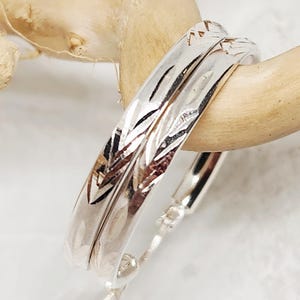 May include: A pair of silver hoop earrings featuring a polished finish and a textured, etched leaf-like pattern along the outer surface. The earrings have a hinged closure mechanism and a rounded, circular shape, reflecting light across the metallic surface.