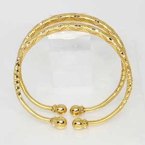 Op de afbeelding: Twee gouden armbanden met een diamantpatroon. De armbanden zijn open en hebben een afgerond uiteinde.