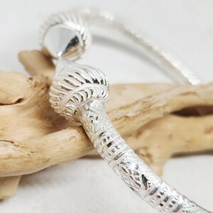 May include: A silver bangle bracelet featuring a textured, engraved surface with intricate geometric patterns. The bracelet ends in two prominent, rounded, ribbed finials that taper to a point, creating a distinct and detailed metallic accessory.