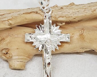 May include: A silver-colored pendant in the shape of a cross, featuring a detailed depiction of Jesus Christ. The cross has a sunburst design around the center and a textured surface. The pendant is suspended from a small loop.