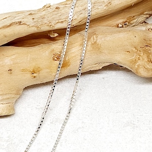 Puede incluir: Primer plano de dos delicadas cadenas de plata. Las cadenas son finas y rectangulares, reflejando la luz. El fondo presenta un trozo de madera flotante marrón claro sobre una superficie blanca. Joyería.