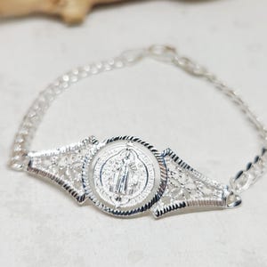 May include: A silver bracelet featuring a detailed medallion with a religious figure. The bracelet has a delicate chain and ornate filigree accents on either side of the medallion. The overall design is intricate and elegant.